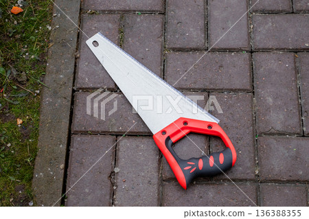 Hand saw with red and black handle on a brick path, detail of a sharp blade and teeth for cutting, gardening tool. Hand saw with red and black handle on a brick path, detail of a sharp blade and teeth for cutting, gardening tool. 136388355