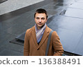 Young Caucasian man in brown coat with a gray bag standing on a wet street. Daily commute or urban lifestyle. Professional lawyer or business manager portrait. 136388491