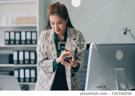 Determined asian businesswoman works late in a modern office, confidently analyzing data on her laptop, embodying success and professionalism with a smile 136388898