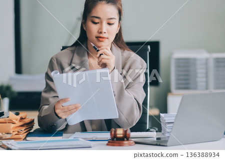 Confident Lawyer Working at Desk: A young Asian female lawyer confidently works on her laptop, surrounded by legal symbols. She exudes professionalism and competence in her modern office setting. 136388934