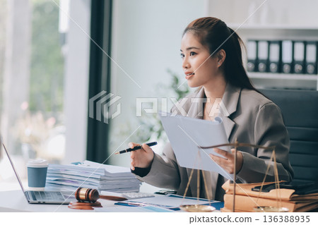 Confident Lawyer Working at Desk: A young Asian female lawyer confidently works on her laptop, surrounded by legal symbols. She exudes professionalism and competence in her modern office setting. 136388935