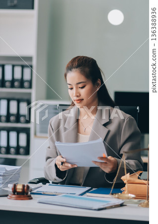 Confident Lawyer Working at Desk: A young Asian female lawyer confidently works on her laptop, surrounded by legal symbols. She exudes professionalism and competence in her modern office setting. Confident Lawyer Working at Desk: A young Asian female lawyer confidently works on her laptop, surrounded by legal symbols. She exudes professionalism and competence in her modern office setting. 136388936