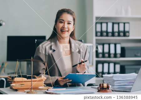 Confident Lawyer Working at Desk: A young Asian female lawyer confidently works on her laptop, surrounded by legal symbols. She exudes professionalism and competence in her modern office setting. Confident Lawyer Working at Desk: A young Asian female lawyer confidently works on her laptop, surrounded by legal symbols. She exudes professionalism and competence in her modern office setting. 136388945