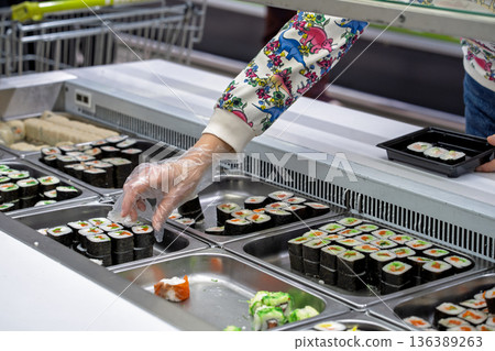 Buying sushi rolls in a store. A customer's hand in a disposable glove selects sushi rolls in a store. Buying sushi rolls in a store. A customer's hand in a disposable glove selects sushi rolls in a store. 136389263
