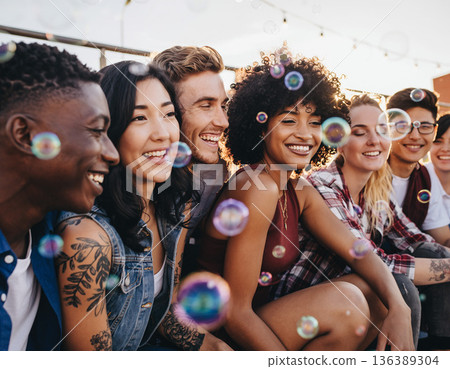 Group of friends enjoying a fun moment together while sitting outdoors in the sunshine with bubbles floating around them Group of friends enjoying a fun moment together while sitting outdoors in the sunshine with bubbles floating around them 136389304
