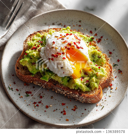Sourdough toast topped with mashed avocado and a poached egg served on a white plate at a kitchen table 136389378