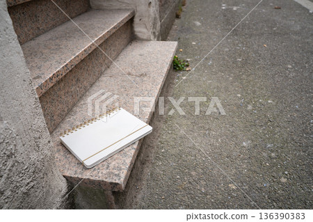 White blank notebook lying on the bottom step outside. Everyday urban scene showing a personal object, representing writing, study, and casual outdoor life. 136390383