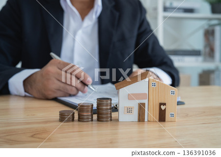 Close-up View of Wooden House Models and Coins on Table with Businessman Writing Notes in Background in Financial Planning Concept 136391036