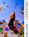 The woman kneels among balloons and toys and blows soap bubbles. 136391487