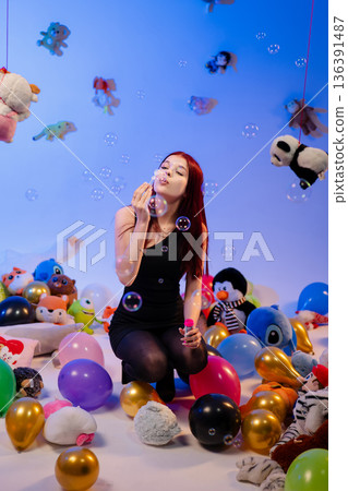 The woman kneels among balloons and toys and blows soap bubbles. 136391487