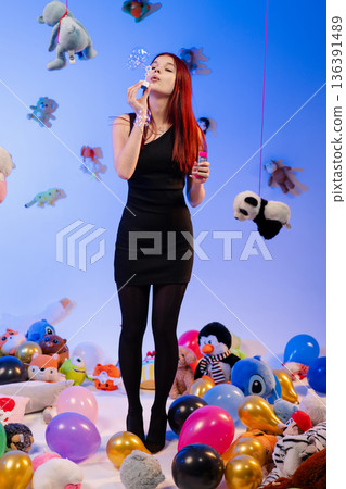 The young woman in a black dress blows soap bubbles among toys and balloons. 136391489