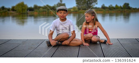 A group of cute little children sit on a wooden pier by a lake. Small children looking into the future. Children sit on a dock. Siblings. Two children of different ages a school-age boy and a 136391624