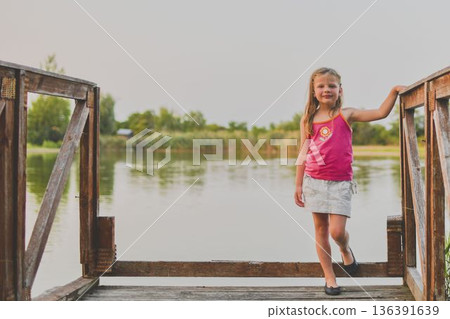 A small smiling girl stands on a wooden pier at the side of a lake. The photo was taken during the golden hour, at sunset. It evokes the concept of childhood, summer vacation and carefree days. A small smiling girl stands on a wooden pier at the side of a lake. The photo was taken during the golden hour, at sunset. It evokes the concept of childhood, summer vacation and carefree days. 136391639