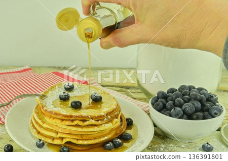 Pouring maple syrup on American pancakes. Sweetened American pancakes with blueberries and maple syrup on a white rustic table. American pancakes on a rustic background. An American classic, this is Pouring maple syrup on American pancakes. Sweetened American pancakes with blueberries and maple syrup on a white rustic table. American pancakes on a rustic background. An American classic, this is 136391801