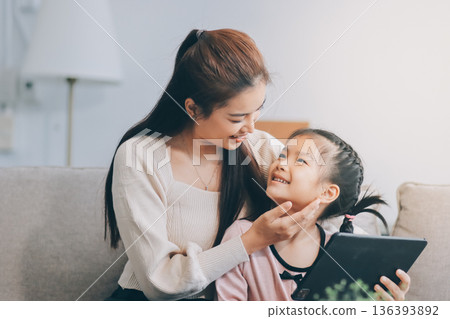 A cheerful Asian mother helps her young daughter do homework and color a book at home, while the father works on a laptop in the background. A cozy, bright, and modern family lifestyle. 136393892