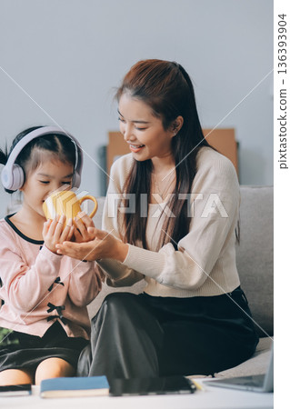 A cheerful Asian mother helps her young daughter do homework and color a book at home, while the father works on a laptop in the background. A cozy, bright, and modern family lifestyle. A cheerful Asian mother helps her young daughter do homework and color a book at home, while the father works on a laptop in the background. A cozy, bright, and modern family lifestyle. 136393904