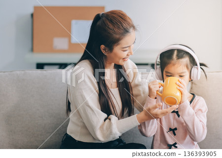 A cheerful Asian mother helps her young daughter do homework and color a book at home, while the father works on a laptop in the background. A cozy, bright, and modern family lifestyle. 136393905