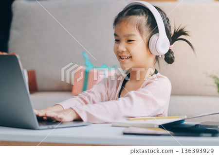 A cheerful Asian mother helps her young daughter do homework and color a book at home, while the father works on a laptop in the background. A cozy, bright, and modern family lifestyle. A cheerful Asian mother helps her young daughter do homework and color a book at home, while the father works on a laptop in the background. A cozy, bright, and modern family lifestyle. 136393929