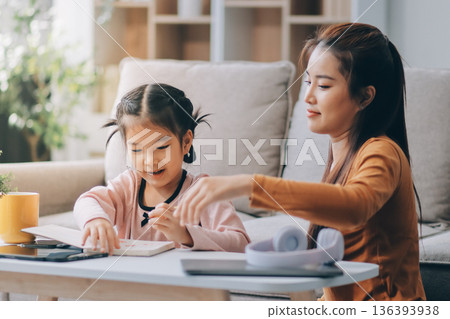 A cheerful Asian mother helps her young daughter do homework and color a book at home, while the father works on a laptop in the background. A cozy, bright, and modern family lifestyle. A cheerful Asian mother helps her young daughter do homework and color a book at home, while the father works on a laptop in the background. A cozy, bright, and modern family lifestyle. 136393938