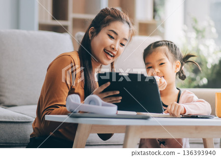 A cheerful Asian mother helps her young daughter do homework and color a book at home, while the father works on a laptop in the background. A cozy, bright, and modern family lifestyle. 136393940