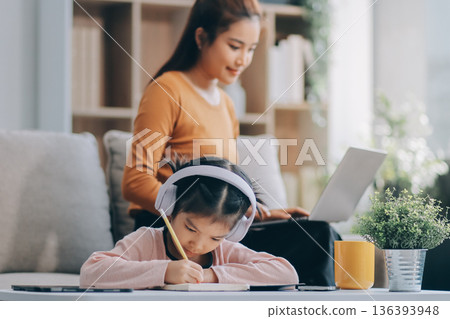 A cheerful Asian mother helps her young daughter do homework and color a book at home, while the father works on a laptop in the background. A cozy, bright, and modern family lifestyle. A cheerful Asian mother helps her young daughter do homework and color a book at home, while the father works on a laptop in the background. A cozy, bright, and modern family lifestyle. 136393948