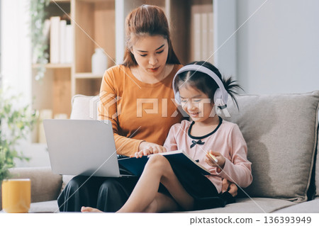 A cheerful Asian mother helps her young daughter do homework and color a book at home, while the father works on a laptop in the background. A cozy, bright, and modern family lifestyle. A cheerful Asian mother helps her young daughter do homework and color a book at home, while the father works on a laptop in the background. A cozy, bright, and modern family lifestyle. 136393949