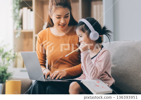 A cheerful Asian mother helps her young daughter do homework and color a book at home, while the father works on a laptop in the background. A cozy, bright, and modern family lifestyle. 136393951