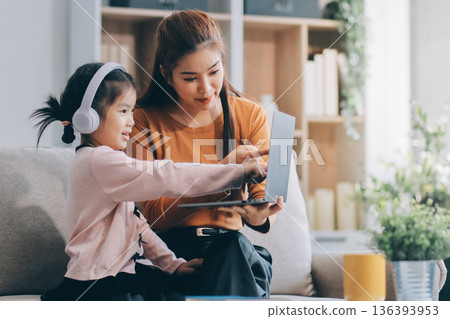A cheerful Asian mother helps her young daughter do homework and color a book at home, while the father works on a laptop in the background. A cozy, bright, and modern family lifestyle. A cheerful Asian mother helps her young daughter do homework and color a book at home, while the father works on a laptop in the background. A cozy, bright, and modern family lifestyle. 136393953