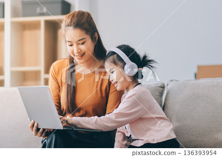 A cheerful Asian mother helps her young daughter do homework and color a book at home, while the father works on a laptop in the background. A cozy, bright, and modern family lifestyle. 136393954