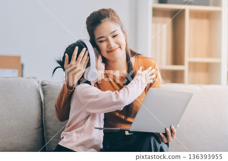 A cheerful Asian mother helps her young daughter do homework and color a book at home, while the father works on a laptop in the background. A cozy, bright, and modern family lifestyle. 136393955