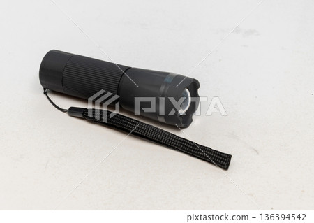 Black portable flashlight with strap lies on a light isolated background. Black portable flashlight with strap lies on a light isolated background. 136394542