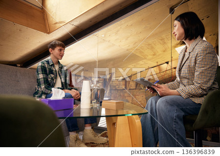 Teenage boy and female psychologist facing each other in counseling room. Teenage boy and female psychologist facing each other in counseling room. 136396839