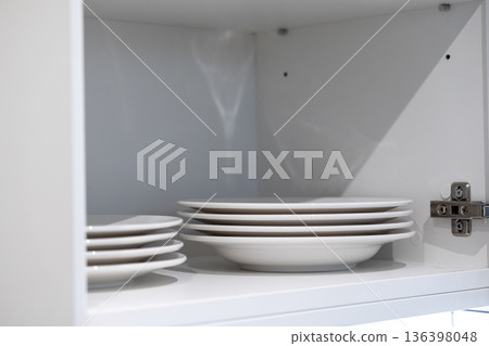 White ceramic plates neatly stacked inside a clean white kitchen cabinet, showcasing minimalist organization and essential domestic kitchenware for an inviting home White ceramic plates neatly stacked inside a clean white kitchen cabinet, showcasing minimalist organization and essential domestic kitchenware for an inviting home 136398048