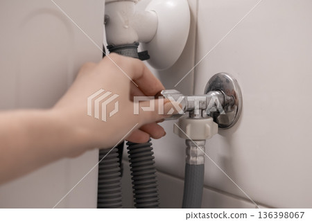 Hand adjusts a chrome water shut-off valve connected to a washing machine hose on a white wall, ensuring proper water supply for home appliance maintenance and plumbing 136398067