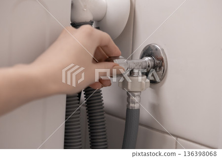 Person's hand turning a chrome water shut-off valve, controlling the water supply to a home appliance during plumbing work or installation 136398068