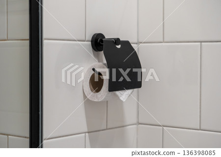 A fresh toilet paper roll on a sleek black holder, neatly positioned on a modern white tiled bathroom wall, emphasizing cleanliness and daily hygiene needs 136398085
