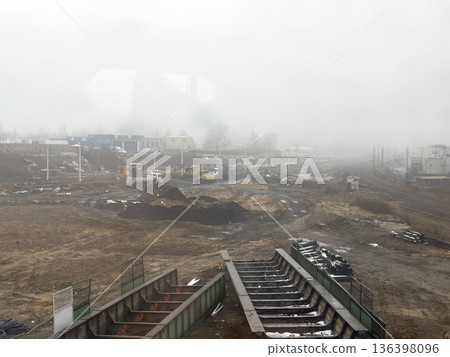 Industrial construction site with heavy equipment, temporary buildings, and distant railroad tracks, obscured by dense fog, representing infrastructure progress Industrial construction site with heavy equipment, temporary buildings, and distant railroad tracks, obscured by dense fog, representing infrastructure progress 136398096