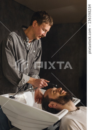 Barber washing client hair with shampoo at salon sink. Professional male grooming service, relaxing head wash process, close up of hairstyling care in modern barbershop. 136398284