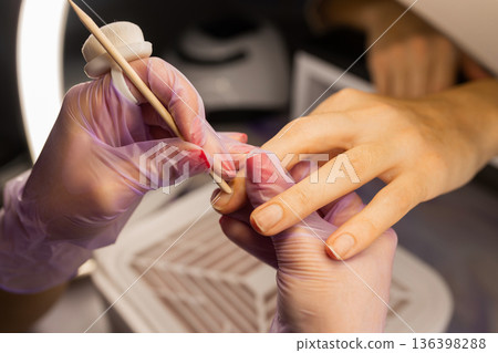 Close up of professional manicure cuticle care using wooden stick and brush. Nail technician wearing gloves cleaning female nails in beauty salon. Nail care, hygiene and cosmetic treatment concept. 136398288