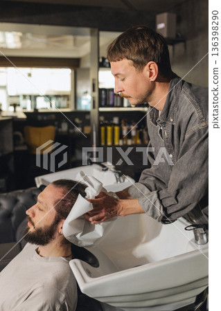 Barber washing client hair with shampoo at salon sink. Professional male grooming service, relaxing head wash process, close up of hairstyling care in modern barbershop. 136398290