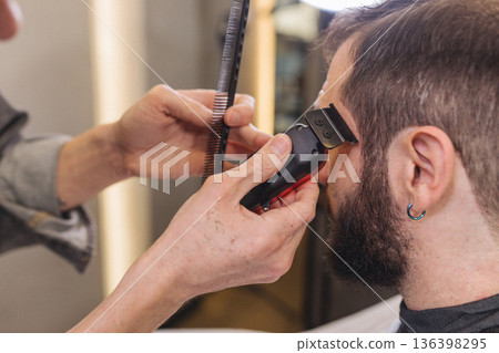 Barber trimming beard of male client with electric clipper in barbershop. Professional grooming process, beard shaping, men haircare routine, classic barber service. 136398295