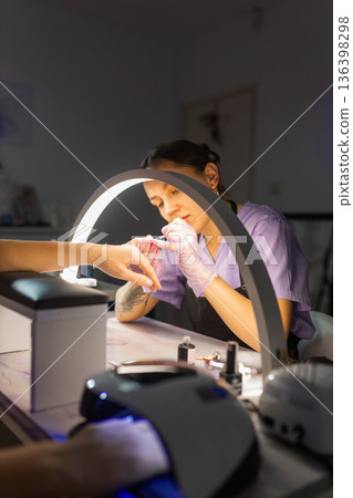 Professional nail technician performing manicure for female client under lamp in beauty salon. Nail care service, cosmetic treatment, hygiene, modern manicure workspace concept. 136398298