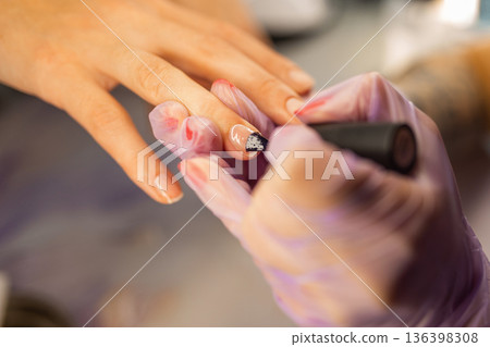 Close up of professional manicure process. Nail technician applying gel polish on female nails with brush while wearing protective gloves in beauty salon Close up of professional manicure process. Nail technician applying gel polish on female nails with brush while wearing protective gloves in beauty salon 136398308