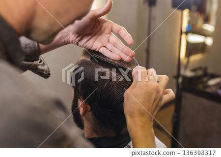Barber cutting male client hair with scissors and comb. Close up of professional haircut process, precision technique, men grooming service in modern barbershop. Barber cutting male client hair with scissors and comb. Close up of professional haircut process, precision technique, men grooming service in modern barbershop. 136398317