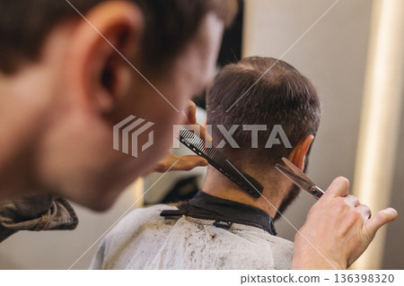 Barber cutting male client hair with scissors and comb. Close up of professional haircut process, precision technique, men grooming service in modern barbershop. Barber cutting male client hair with scissors and comb. Close up of professional haircut process, precision technique, men grooming service in modern barbershop. 136398320