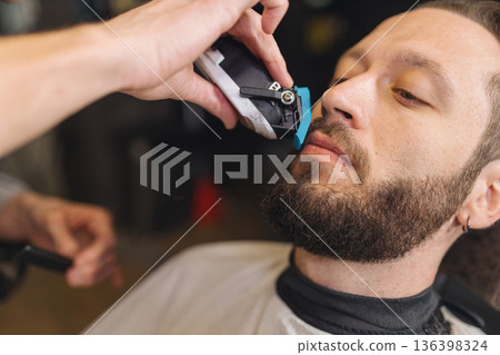 Barber trimming beard of male client with electric clipper in barbershop. Professional grooming process, beard shaping, men haircare routine, classic barber service. Barber trimming beard of male client with electric clipper in barbershop. Professional grooming process, beard shaping, men haircare routine, classic barber service. 136398324