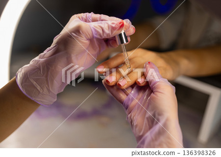 Close up of professional manicure care with cuticle oil application. Nail technician wearing gloves applying nourishing serum on female nails in beauty salon. Close up of professional manicure care with cuticle oil application. Nail technician wearing gloves applying nourishing serum on female nails in beauty salon. 136398326