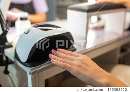 Close up of female hand curing gel nail polish under UV LED lamp in beauty salon. Professional manicure process, nail drying, cosmetic treatment, hygiene and nail care concept. 136398330