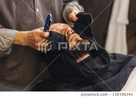 Relaxed male client lying in barber chair during classic shaving procedure. Professional barbershop service, beard grooming, comfort and men self care lifestyle concept. Relaxed male client lying in barber chair during classic shaving procedure. Professional barbershop service, beard grooming, comfort and men self care lifestyle concept. 136398344