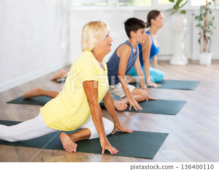 Old woman practicing pigeon pose of yoga in light fitness room 136400110